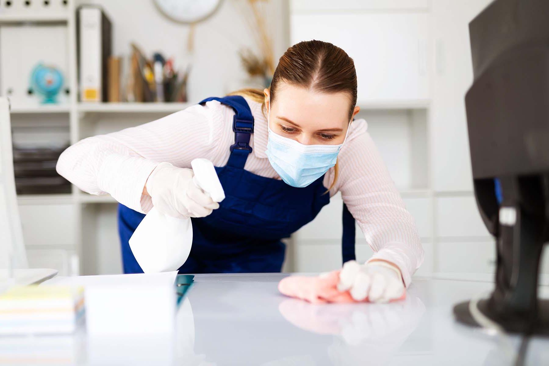 Woman in a mask sanitizing an office desk.