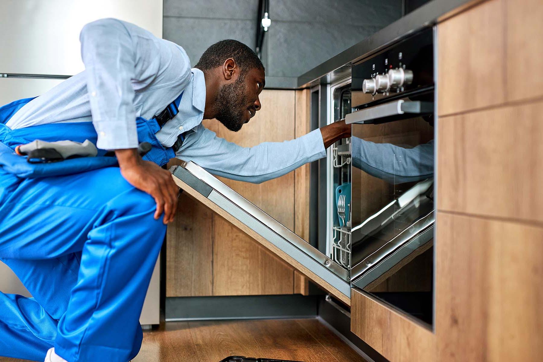 A technician in blue overalls inspects a dishwasher.