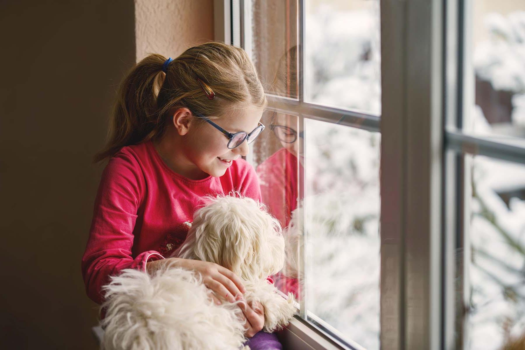 戴眼镜的女孩抱着一只狗，望着白雪覆盖的窗外.